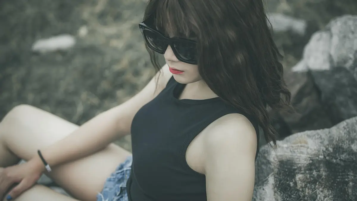 A woman wearing a tank top, shorts and sunglasses by rocks.